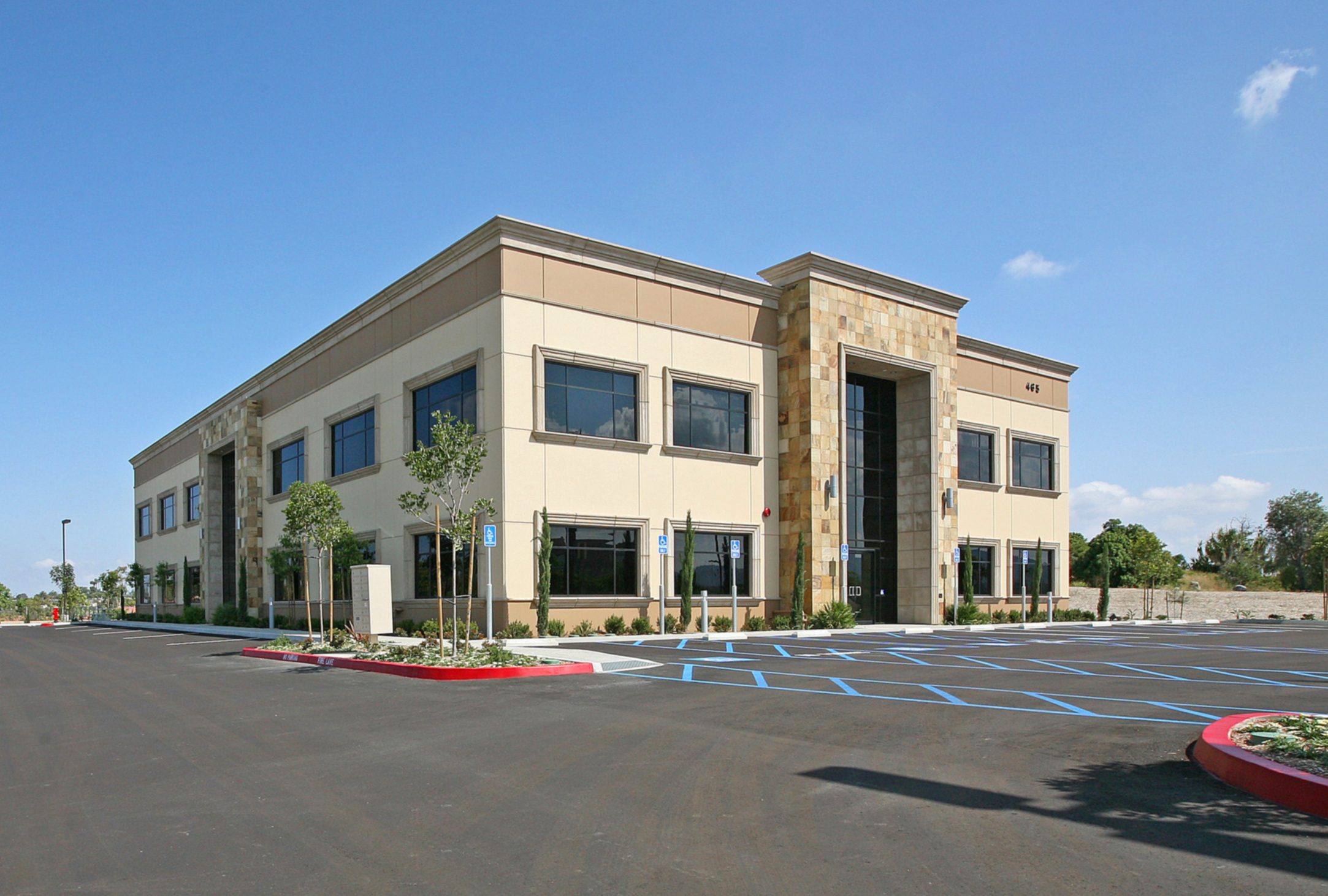 Hacienda+Photo+1a A two-story commercial building with large windows, beige exterior, and a central entrance, surrounded by an empty parking area under a clear blue sky.