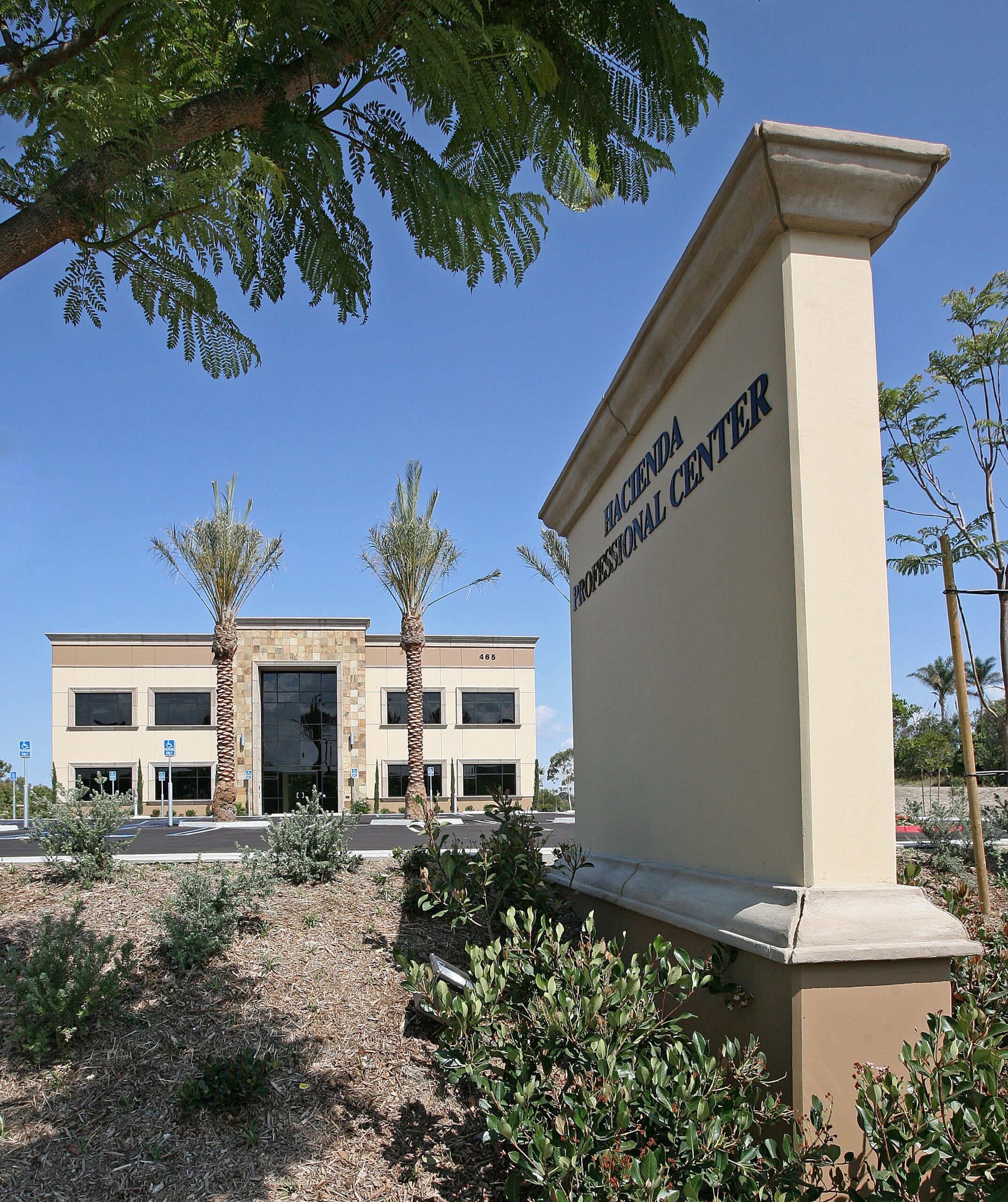 IMG_2983 A beige sign reading "Hacienda Professional Center" stands in front of a two-story office building with palm trees and landscaped grounds under a clear blue sky.