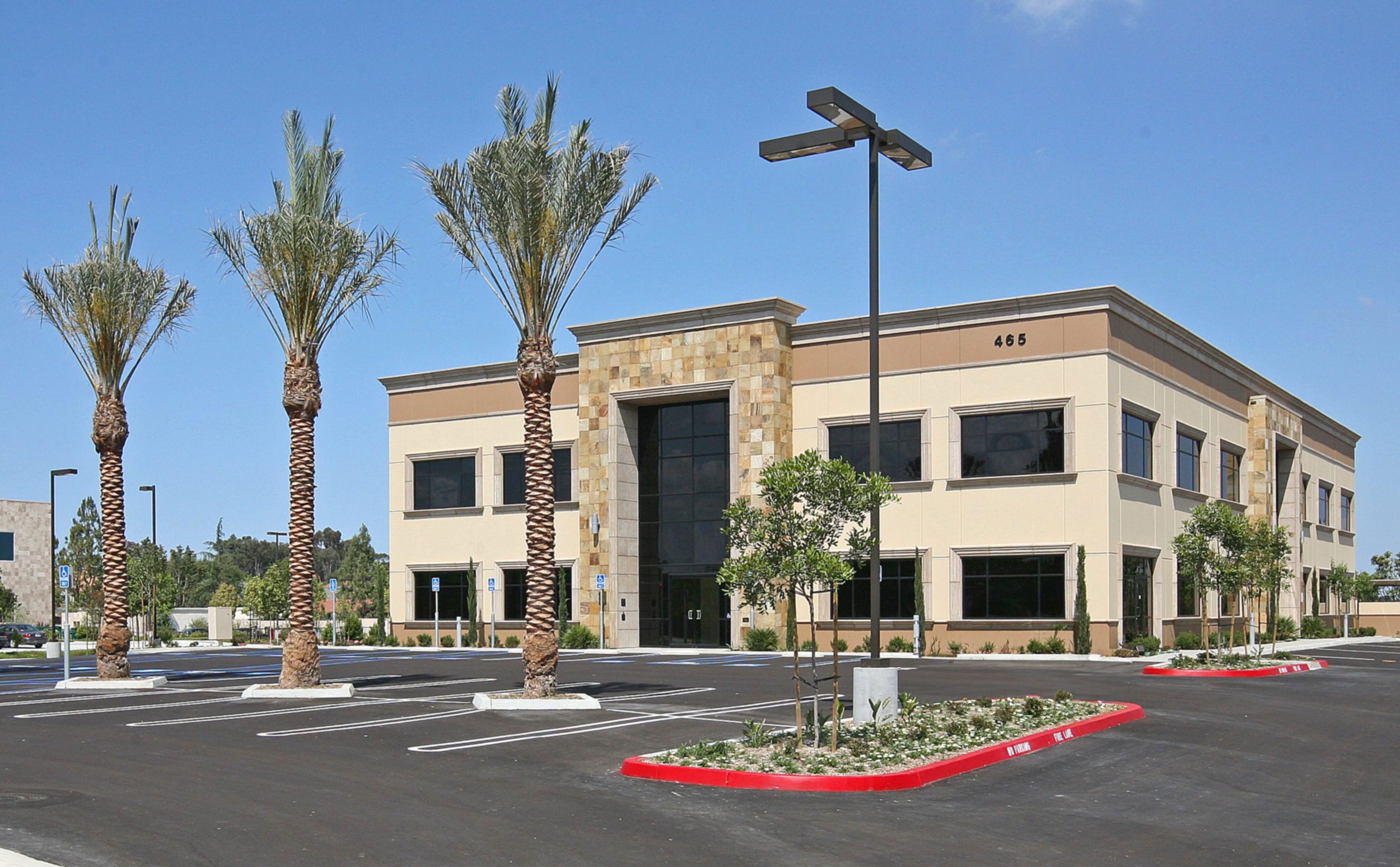 IMG_3005cropped-img-ps A two-story commercial building with large windows and stone accents, surrounded by an empty parking lot with palm trees and small plants. The building number "465" is visible.